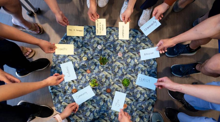 A circle of hands reach toward the center, each holding a piece of paper with positive phrases on them, such as "joy", "community", "family", and "pay it forward."